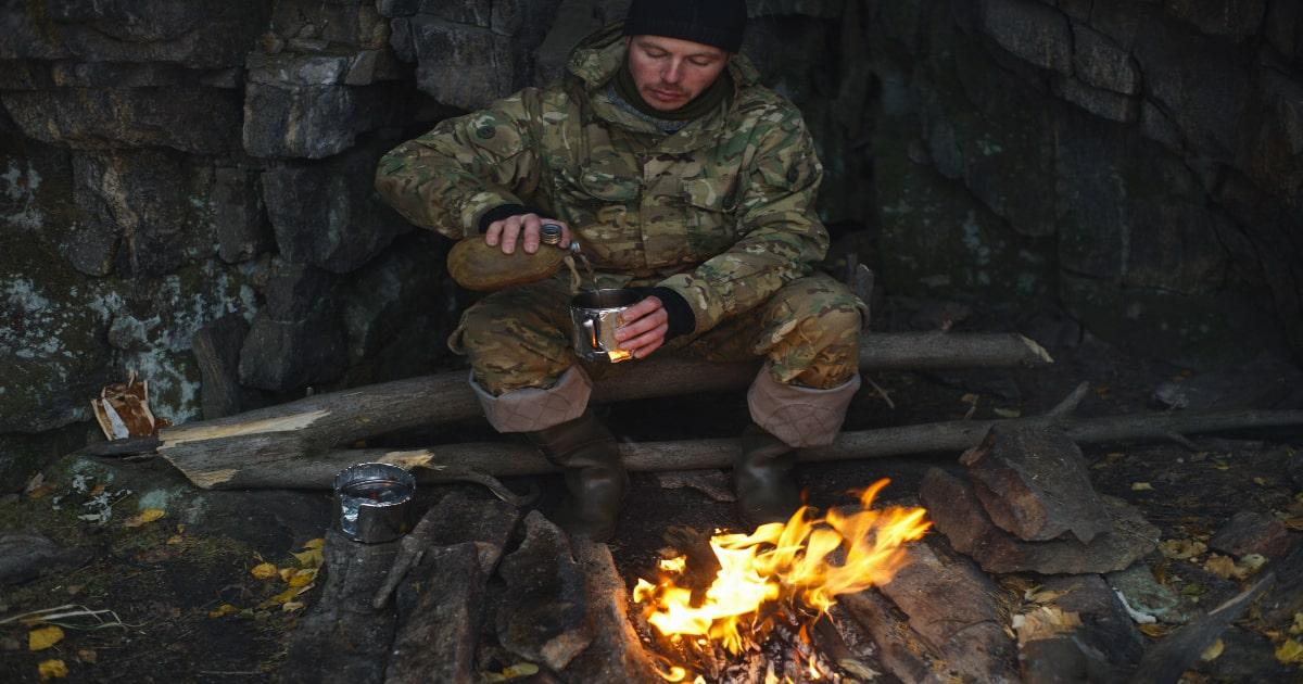 man in camouflage by fire pouring warm drink